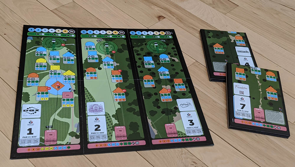 The image shows components of a disc golf  board game including cards, player boards, and tokens laid out on a wooden floor.
