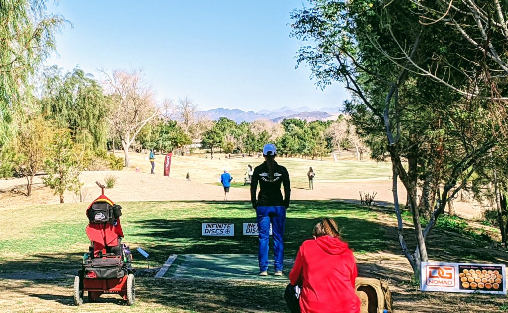 A group of people are enjoying a sunny day at a park with disc golf equipment and a mountainous backdrop.
